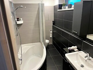 Moderní koupelna s dlažbou, rohovou vanou a tmavými obklady, nabízí světlý a elegantní design.