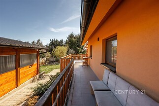 Prostorná terasa s dlažbou a dřevěným zábradlím, vhodná k relaxaci. Nabízí výhled do zahrady a přístup do přízemního domu s moderní fasádou.