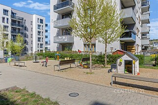 Dětské hřiště v moderním bytovém komplexu se zpevněnými cestami, lavičkami a herními prvky. Okolí dominuje moderní architektura a upravená zeleň.