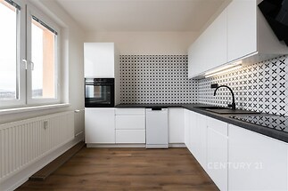 Moderní kuchyň s parketovou podlahou, světlými bílými skříňkami a stylovým keramickým obkladem. Velká okna zajišťují dostatek světla.