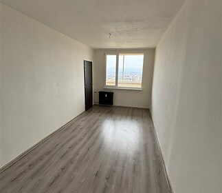 Úzký pokoj s laminátovou podlahou a velkým oknem, výhled na město. Světlý prostor vhodný k rekonstrukci.