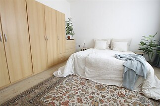 Světlá ložnice s dřevěnými vestavěnými skříněmi, parketovou podlahou a velkým kobercem, moderní design s rostlinami pro útulnou atmosféru.