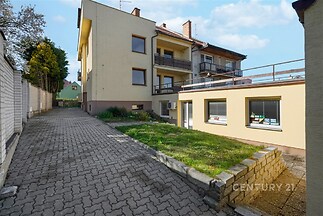 Venkovní prostor s dlážděnou příjezdovou cestou a udržovaným trávníkem, část moderního bytového domu s prostornými balkony a velkými okny.