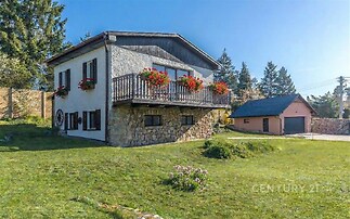 Dvoupodlažní dům na velkém pozemku, obložený kamenem, s květinovými truhlíky na balkoně, zelený a klidný okolní prostor.