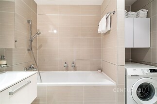 Moderní koupelna s novými obklady, vanou a pračkou. Světlé dlažby a čistý design zvyšují pocit prostornosti a komfortu.