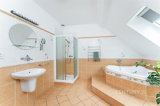 Prostorná koupelna se střešním oknem, dlažba v teplých tónech, moderní sprchový kout a rohová vana, světlý interiér.
