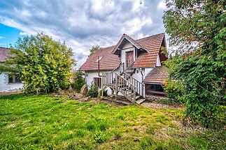 Tradiční venkovský dům se sedlovou střechou a dřevěným schodištěm, obklopený zelenou zahradou, ideální pro klidné bydlení.