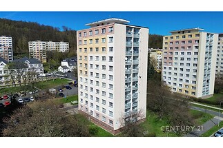 Vyšší bytový dům s barevnou fasádou, obklopen zelení a sousední zástavbou, situovaný v klidné rezidenční čtvrti blízko parku.