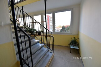Světlé schodiště s velkými okny a koberci, kovové zábradlí, výhled na okolní bytové domy, dobře udržované společné prostory.