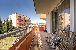 Prostorný balkon s dlažbou a moderním zábradlím, výhled na novostavby a okolní zeleň, ideální pro relaxaci a venkovní posezení.