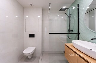 Moderní koupelna, světlé dlaždice, prosklený sprchový kout, dřevěný nábytek, závěsné WC, elegantní design.