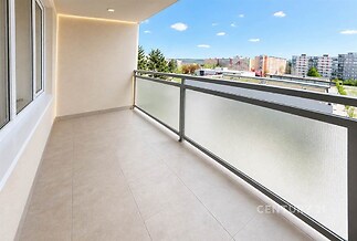Prostorný balkon s dlažbou, moderním zábradlím a velkými okny poskytuje světlé a vzdušné prostředí s výhledem na městskou zástavbu