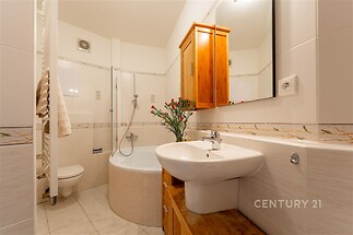 Koupelna s dlažbou, rohovou vanou a dřevěnou skříňkou, moderní styl s velkým zrcadlem a květinovou dekorací.