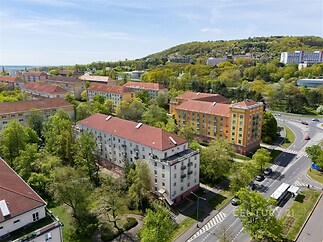 Aerial view of a residential area with colorful apartment buildings surrounded by greenery, a curving road, and distant hills in the background.