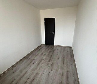 Úzký pokoj s moderní parketovou podlahou, bílými stěnami a tmavými dveřmi, vhodný pro vestavěné skříně nebo úložný prostor.