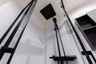 Moderní sprchový kout s černými skleněnými dveřmi, elegantním dešťovým sprchovým panelem a světlými keramickými obklady