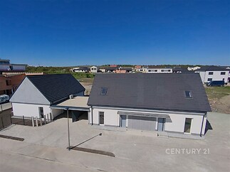 Moderní rodinný dům s tmavou střechou a bílou fasádou, terasa z dlažby, nachází se na klidném předměstí s otevřeným prostorem kolem.