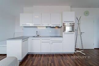 Moderní kuchyň s bílými lesklými skříňkami, vestavěnou troubou a parketovou podlahou, prostorný design pro pohodlné bydlení.