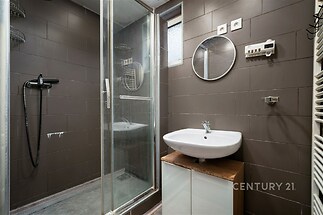 Moderní koupelna s tmavou dlažbou, sprchovým koutem a kulatým zrcadlem. Elegantní design s bílým umyvadlem na dřevěné skříňce.