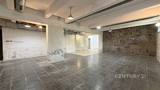 Průmyslový prostor s betonovými sloupy, dlažbou a kamennou zdí. Vysoký strop a moderní osvětlení poskytují industriální atmosféru.