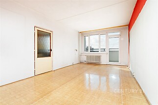 Světlý obývací pokoj s parketovou podlahou, velká okna poskytující výhled, moderní stav, radiátor pod okny pro vytápění.