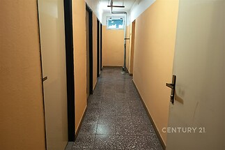 Úzká chodba s dlažbou, jednoduchými dveřmi a malým oknem, v udržovaném stavu, vhodná pro bytové jednotky nebo kancelářské prostory.