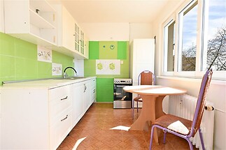 Světlá kuchyň s parketovou podlahou, velkými okny a moderními bílými skříňkami, které dodávají prostoru svěží a rekonstruovaný vzhled.