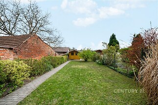 Zahrada s udržovaným trávníkem, dlážděným chodníkem a zahradním domkem, obklopená zelení a cihlovými stavbami, nabízí klidné prostředí.