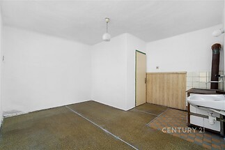 Prázdná místnost s dlažbou a dřevěným obložením, použitá jako kuchyňský prostor. Starší zařízení s umyvadlem a dveřmi k dalšímu vstupu.