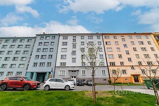 Vícepatrový bytový dům s pastelově barevnou fasádou a parkovacími místy před budovou, nachází se v městské zástavbě.