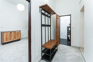 Předsíň s dlažbou, věšákem a dřevěným úložným prostorem, sousedící s moderní kuchyní světlých tónů.