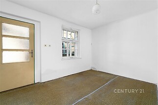 Prázdná místnost s bílými stěnami, světlé okno a starší podlahová krytina, dřevěné dveře, vhodná k rekonstrukci.