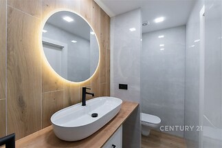 Koupelna s moderním designem, dřevěný obklad, oválné osvětlené zrcadlo a keramická umyvadlová deska vytvářejí elegantní vzhled.