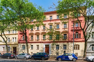 Historická fasáda bytového domu s barevným omítkovým stylem, renovovaná se zdobenými okny, před domem stromy a zaparkovaná auta