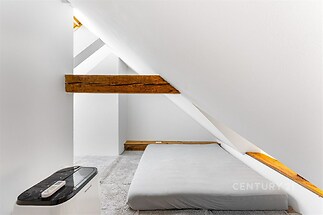 Podkrovní ložnice s dřevěnými trámy, světlé stěny a šedý koberec, moderní minimalistický design, vytváří útulnou atmosféru.