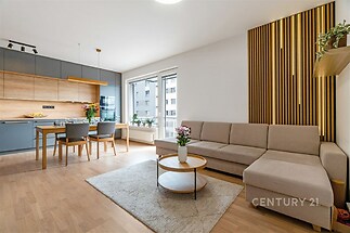 Světlý obývací pokoj s parketovou podlahou, velkými okny a moderní kuchyní. Pohodlná rohová sedačka a jídelní stůl s dřevěným nábytkem.