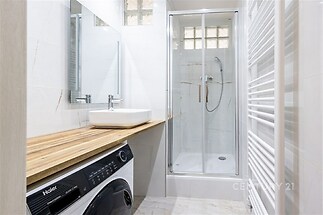 Moderní koupelna se sprchovým koutem, dřevěnou deskou a světlou dlažbou. Pračka s horním plněním a minimalistický styl přinášejí praktičnost.