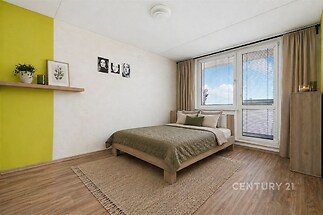 Světlá ložnice s parketovou podlahou, velká okna pro přirozené světlo, moderní nábytek a dekorativní prvek na stěně.