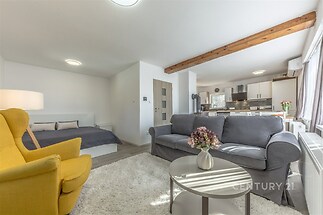 Světlý obývací pokoj s parketovou podlahou a velkými okny, moderní styl s dřevěnými prvky a pohodlným nábytkem, propojený s kuchyní.