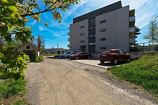 Moderní bytový dům s parkováním na venkovním parkovišti, obklopený zelení. Budova má rovnou fasádu a balkony, což dodává moderní vzhled.