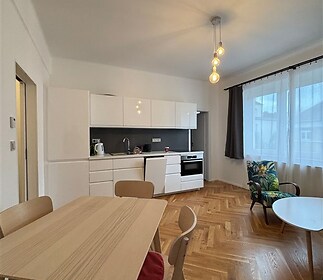 Kuchyňský kout s moderními bílými skříněmi a parketovou podlahou, prostorné dispozice, velká okna poskytující dostatek denního světla.