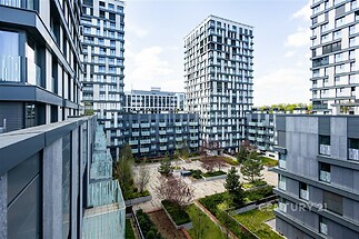 Výhled z balkonu na moderní bytový komplex s upraveným vnitroblokem, velkými okny a šedou fasádou.