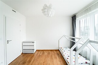 Dětský pokoj s parketovou podlahou, velkými okny a moderním designem, vybavený jednolůžkovou postelí a bílým úložným nábytkem.