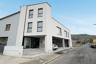 Moderní dvoupatrový dům s šedou fasádou, dlažbou u vchodu a parkovacími místy. Nachází se v klidné rezidenční čtvrti.