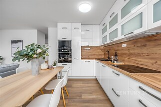 Moderní kuchyň s dřevěnou pracovní deskou, vestavěnými spotřebiči a parketovou podlahou. Světlý interiér s bílými skříňkami