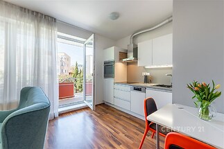 Moderní kuchyň s parketovou podlahou, integrovanými spotřebiči a velkými okny, které poskytují přístup na balkon, působí světlým dojmem.