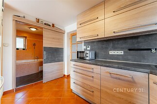 Moderní kuchyň se světlými dřevěnými skříňkami, dlažbou a velkými úložnými prostory, sousedící s koupelnou s velkou vanou.