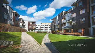 Moderní bytový komplex s udržovaným trávníkem, dlážděnými chodníky a velkými okny. Architektura kombinuje dřevo a beton pro moderní vzhled.