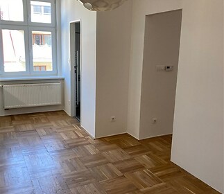 Světlý obývací pokoj s parketovou podlahou a velkými okny, moderní svítidlo, čisté bílé stěny v rekonstruovaném bytě.