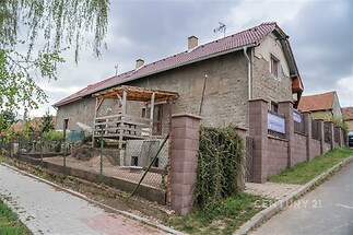 Kamenný rodinný dům s šikmou střechou a velkou terasou, obklopený cihlovým plotem a zahradou, nachází se v klidné vesnické lokalitě.
